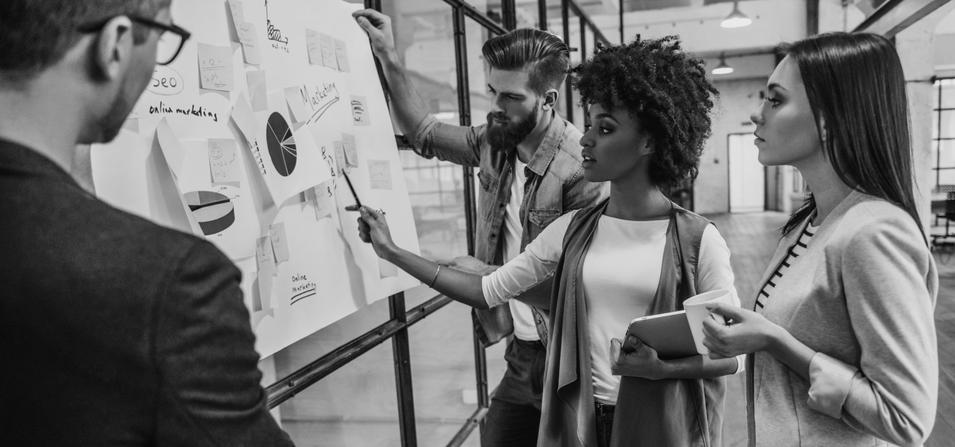 Collaboration is a key to best results. Group of young modern people in smart casual wear planning business strategy while young woman pointing at infographic displayed on the glass wall in the office